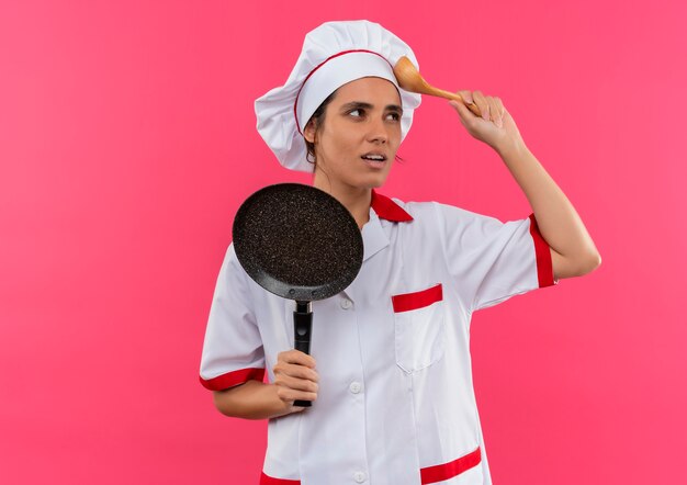 Penser jeune femme cuisinier portant un uniforme de chef tenant une poêle à frire mettant une cuillère sur le front avec copie espace