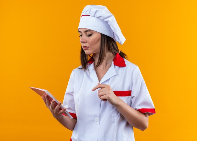 Penser jeune femme cuisinier portant l'uniforme de chef tenant et regardant un ordinateur portable isolé sur fond orange