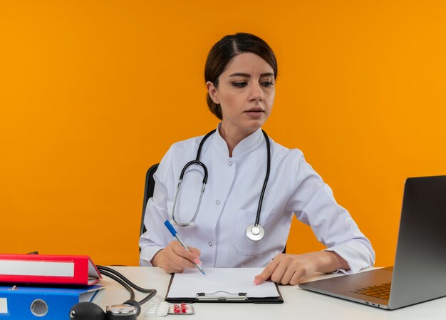 Penser jeune femme médecin portant une robe médicale avec stéthoscope assis au bureau de travail sur ordinateur avec des outils médicaux à la recherche d'un ordinateur portable sur fond jaune d'isolement