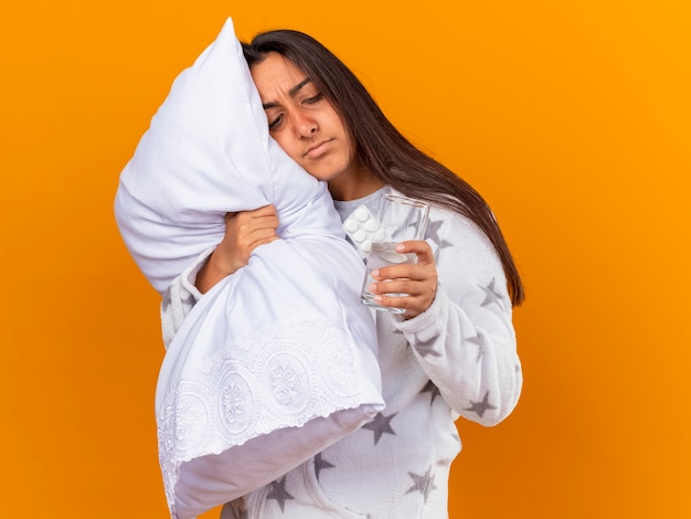 Penser jeune fille malade hugging oreiller tenant et regardant des pilules avec un verre d'eau isolé sur fond jaune