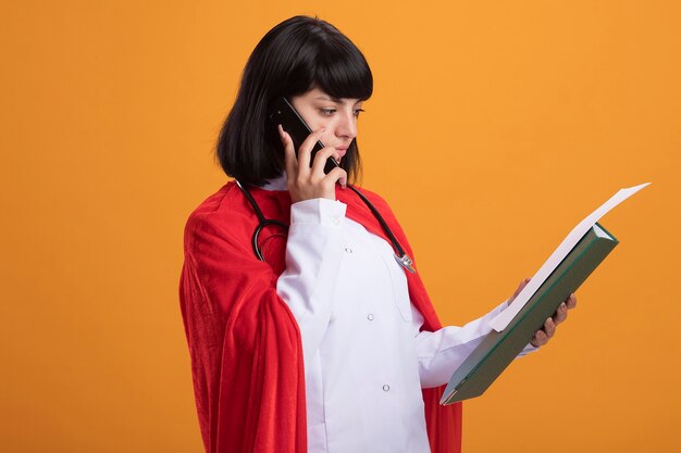 Penser jeune fille de super-héros portant un stéthoscope avec une robe médicale et une cape parle au téléphone et en regardant du papier dans sa main isolé sur un mur orange