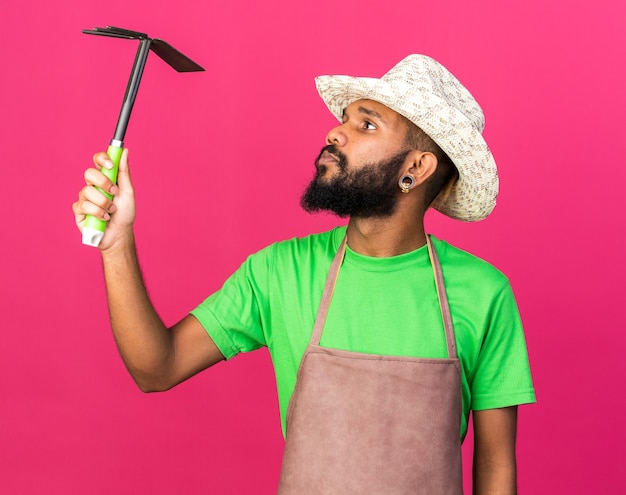 Photo gratuite penser le jeune homme afro-américain de jardinier portant le chapeau de jardinage tenant et regardant le râteau de houe