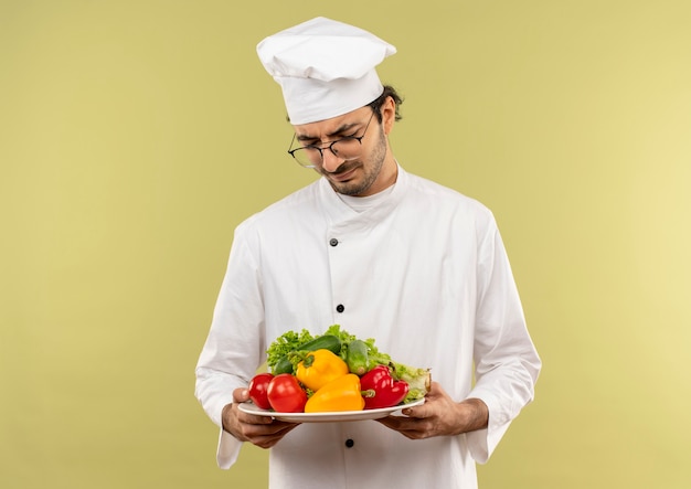 Penser jeune homme cuisinier portant l'uniforme de chef et des lunettes tenant et à la recherche de légumes sur une plaque isolée sur un mur vert