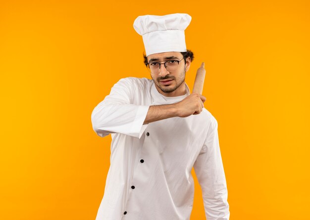 Penser jeune homme cuisinier portant l'uniforme de chef et des lunettes tenant un rouleau à pâtisserie autour de l'épaule isolé sur mur jaune