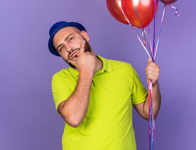 Penser un jeune homme portant un chapeau de fête tenant des ballons et attrapé le menton