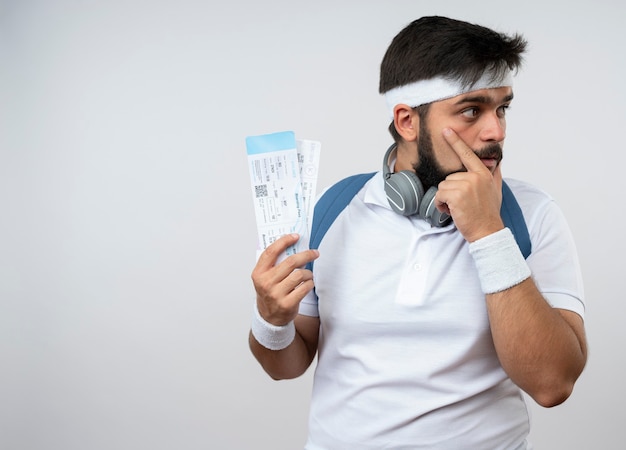 Penser jeune homme sportif regardant côté portant bandeau et bracelet avec sac à dos tenant des billets mettant la main sur le visage isolé sur un mur blanc avec espace copie