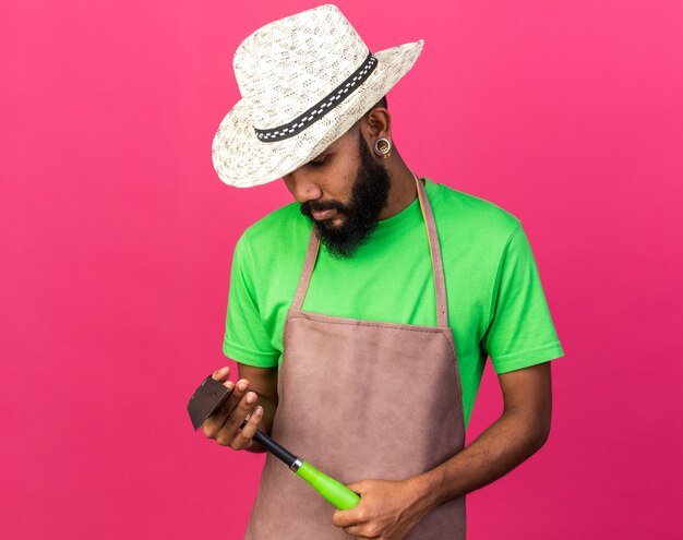 Penser un jeune jardinier afro-américain portant un chapeau de jardinage tenant et regardant un râteau de houe isolé sur un mur rose