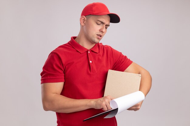 Penser jeune livreur en uniforme avec capuchon tenant la boîte et regardant le presse-papiers dans sa main isolé sur mur blanc