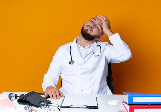 Penser jeune médecin de sexe masculin chauve portant une robe médicale et un stéthoscope assis au bureau de travail avec des outils médicaux mettant la main sur la tête isolée sur un mur orange
