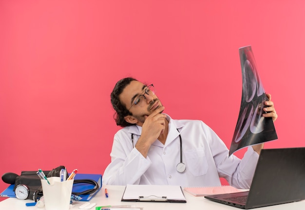 Penser un jeune médecin de sexe masculin avec des lunettes médicales portant une robe médicale avec un stéthoscope assis au bureau