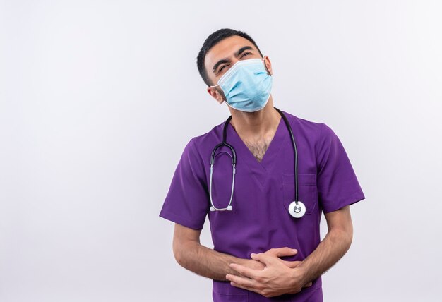 Penser jeune médecin de sexe masculin portant des vêtements de chirurgien violet et un masque médical stéthoscope a mis ses mains sur le ventre sur blanc isolé