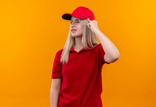 Penser la livraison jeune fille portant un t-shirt rouge et une casquette en orthèse dentaire a mis son doigt sur la tête sur un mur orange isolé