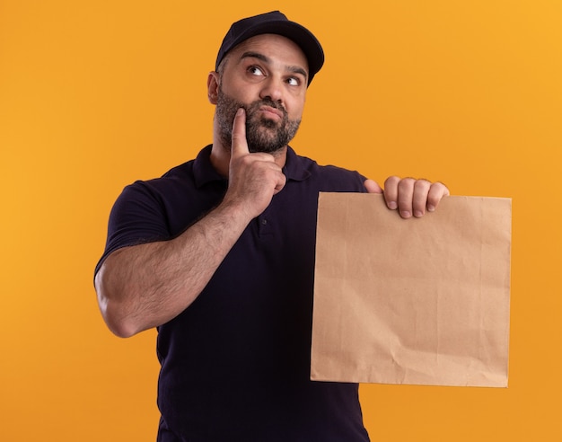 Penser le livreur d'âge moyen en uniforme et cap tenant le paquet alimentaire papier mettant le doigt sur la joue isolé sur mur jaune