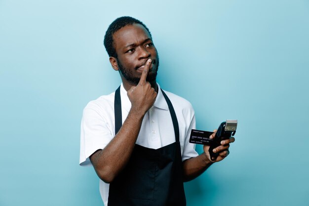 Penser mettre le doigt sur la bouche tenant la carte avec des tondeuses à cheveux jeune coiffeur afro-américain en uniforme isolé sur fond bleu