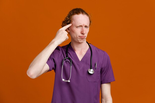 Penser mettre le doigt sur le front jeune médecin de sexe masculin portant l'uniforme avec stéthoscope isolé sur fond orange