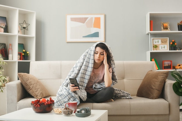 Penser mettre la main sur le temple jeune fille enveloppée dans un plaid tenant et regardant le téléphone assis sur un canapé derrière une table basse dans le salon