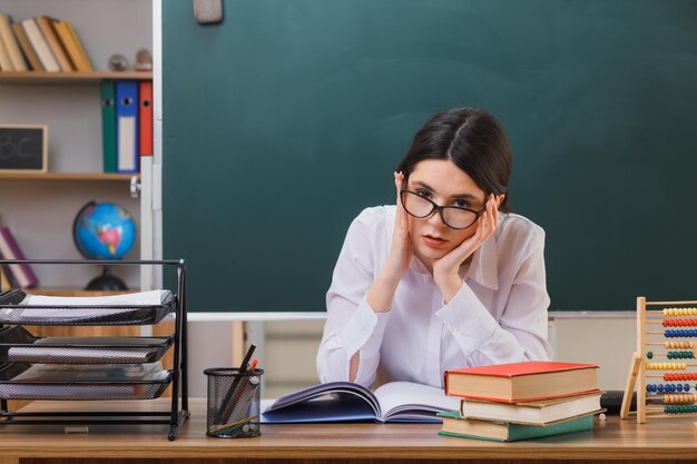 penser mettre les mains sur la joue jeune enseignante portant des lunettes assis au bureau avec des outils scolaires en classe