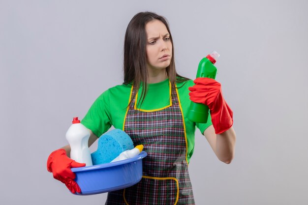 Penser le nettoyage jeune fille portant l'uniforme dans des gants rouges tenant des outils de nettoyage et à la recherche d'un agent de nettoyage sur sa main sur un mur blanc isolé