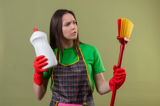 Penser le nettoyage de jeune fille en uniforme dans des gants rouges tenant un agent de nettoyage à la vadrouille sur sa main sur un mur vert isolé