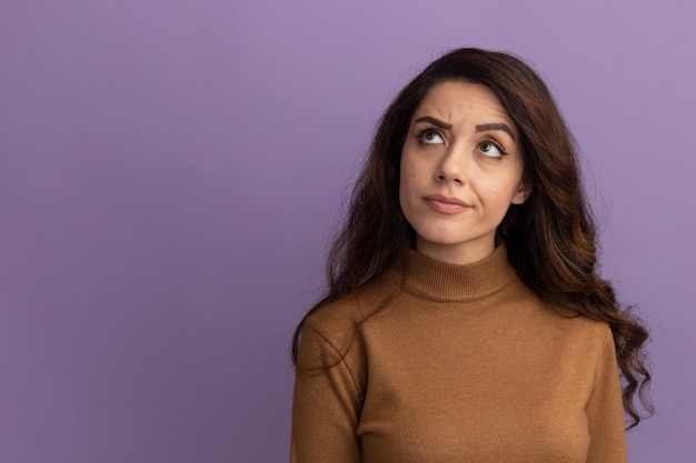 Penser à la recherche d'une belle jeune fille portant un pull à col roulé marron isolé sur un mur violet avec espace de copie