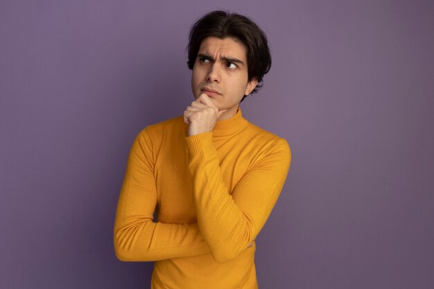 Penser à la recherche de côté jeune beau mec portant un pull à col roulé jaune attrapé le menton isolé sur un mur violet avec copie espace