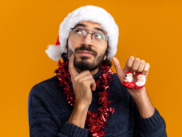 Penser à la recherche de jeune beau mec portant un chapeau de Noël avec guirlande sur le cou tenant des jouets mettant le doigt sur la joue isolé sur fond orange