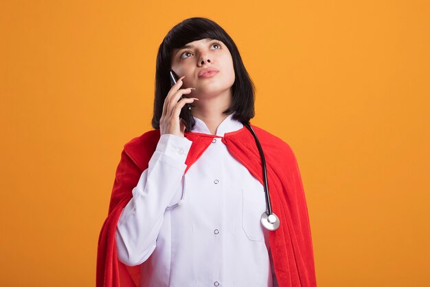 Penser à la recherche de jeune fille de super-héros portant un stéthoscope avec une robe médicale et un manteau parle au téléphone isolé sur un mur orange