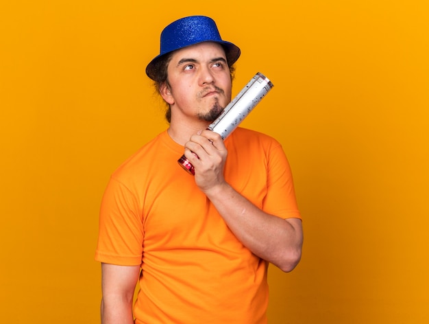 Penser à La Recherche D'un Jeune Homme Portant Un Chapeau De Fête Tenant Un Canon à Confettis Isolé Sur Un Mur Orange
