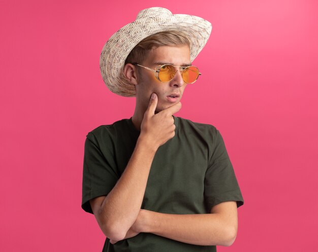 Penser en regardant côté jeune beau mec portant une chemise verte et des lunettes avec un chapeau attrapé le menton isolé sur un mur rose