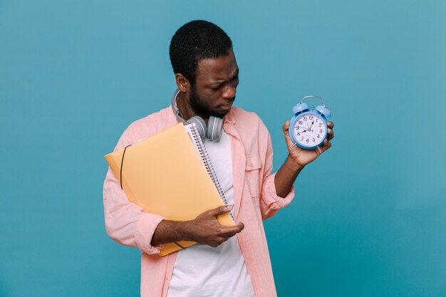 Penser tenant des livres avec réveil jeune homme afro-américain portant des écouteurs sur le cou isolé sur fond bleu