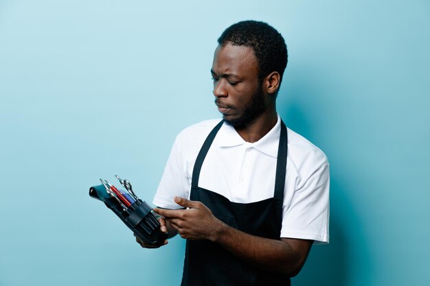 Penser tenant des outils de coiffeur jeune coiffeur afro-américain en uniforme isolé sur fond bleu