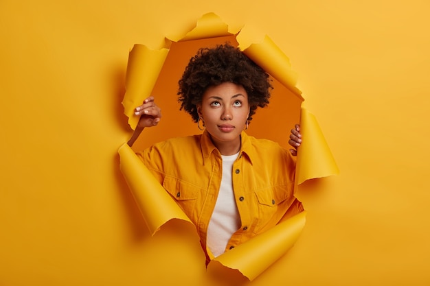 Pensive adorable femme garde le regard vers le haut, se tient dans un trou de papier déchiré, habillée en tenue à la mode, pense à quelque chose