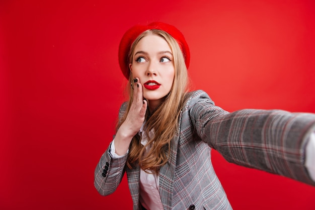 Photo gratuite pensive belle fille en béret prenant selfie. jolie jeune femme en veste élégante posant sur le mur rouge.