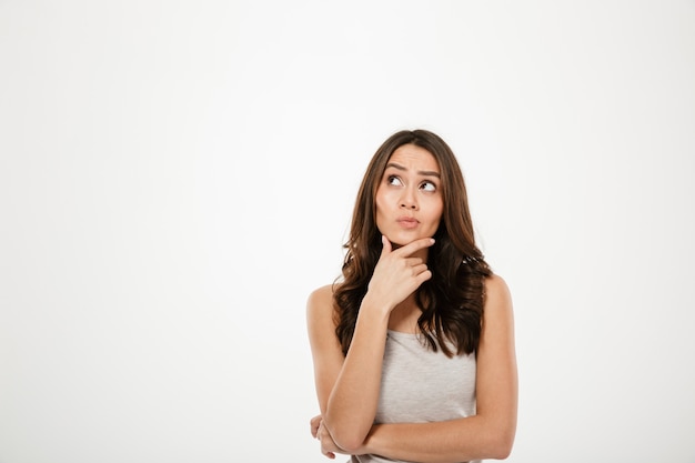 Pensive brunette woman holding her chin and looking up over grey