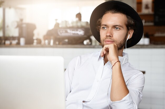 Pensive jeune homme barbu portant une chemise blanche et un chapeau noir touchant son menton et regardant devant lui avec un léger sourire, utilisant un ordinateur portable pour écouter sa musique préférée en ligne dans des écouteurs
