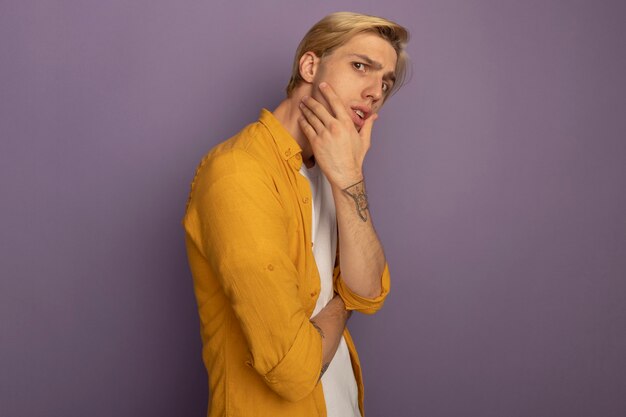 Pensive jeune mec blond portant un t-shirt jaune mettant la main sur la joue isolé sur violet avec copie espace