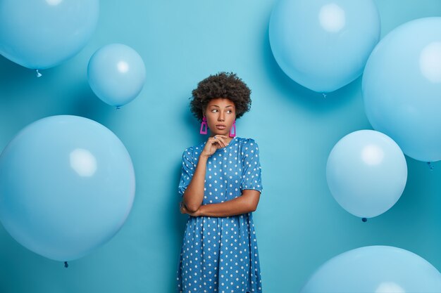 Pensive pensive jeune femme à la peau sombre a les cheveux bouclés, regarde de côté avec une expression de rêve, aime les vacances d'été et la célébration, se tient à l'intérieur, regarde de côté, pose contre le mur bleu