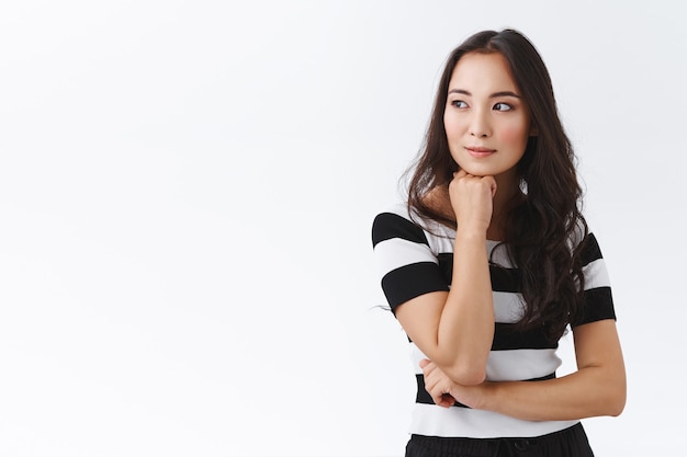 Pensive, séduisante et réfléchie, jeune femme brune asiatique en t-shirt rayé, tenir la main sous le menton, détournant le regard rêveuse, souriante en contemplant quelqu'un qui passe, debout sur fond blanc