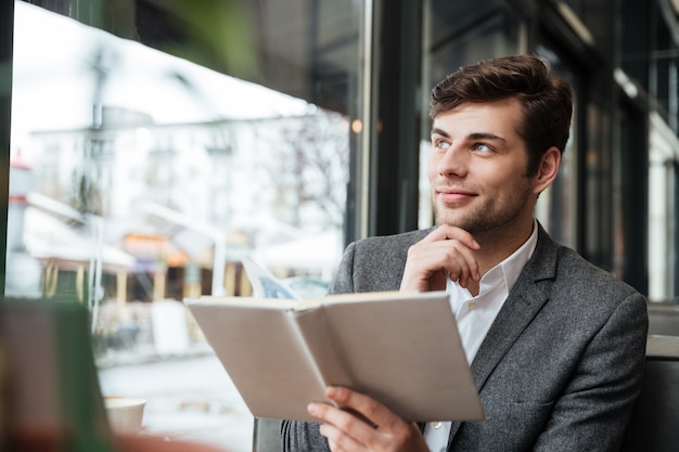 Pensive smiling businessman sitting by the table in cafe with laptop computer while holding book and looking away