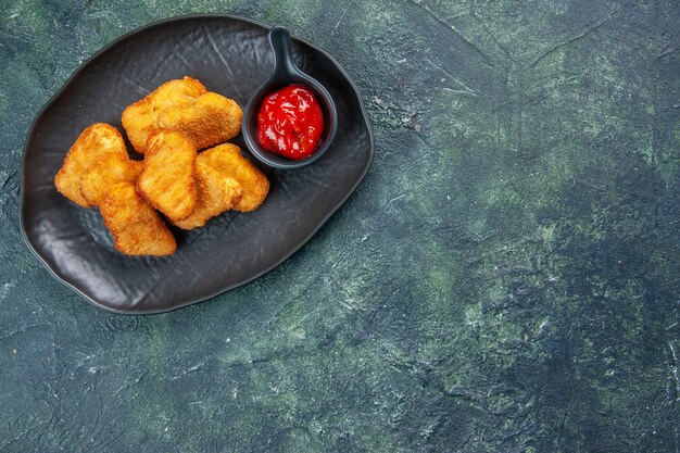 Pépites de poulet savoureuses et ketchup dans des assiettes noires sur le côté droit sur une surface sombre