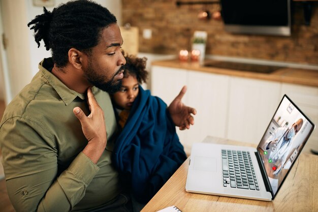Un père afro-américain ayant un appel vidéo avec un médecin de famille pendant que sa fille ne se sent pas bien