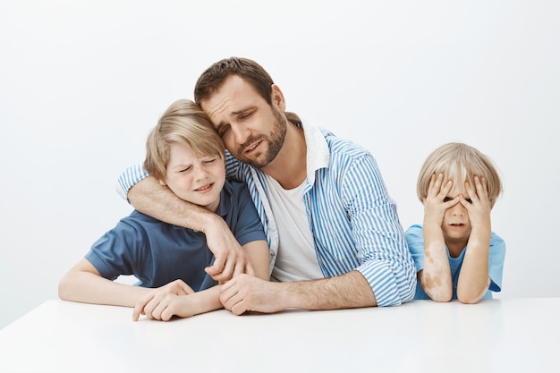 Père chagrin avec ses fils assis à table, étreignant le garçon et pleurant, étant bouleversé et malheureux alors que son plus jeune fils n'a rien à faire