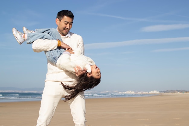 Photo gratuite père et fille ludiques passent du temps sur la plage. famille japonaise marchant, étreignant, riant, s'amusant. loisirs, temps en famille, concept parental