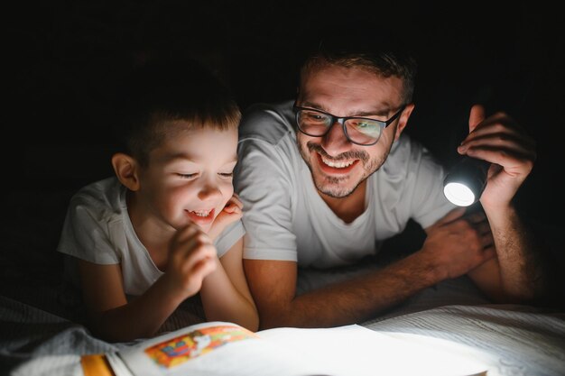 Père et fils avec lampe de poche lisant un livre sous couverture à la maison