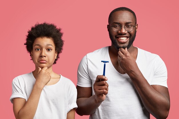 Un père joyeux tient le rasoir, touche sa barbe épaisse, dit des conseils à l'adolescent sur la façon de se raser correctement