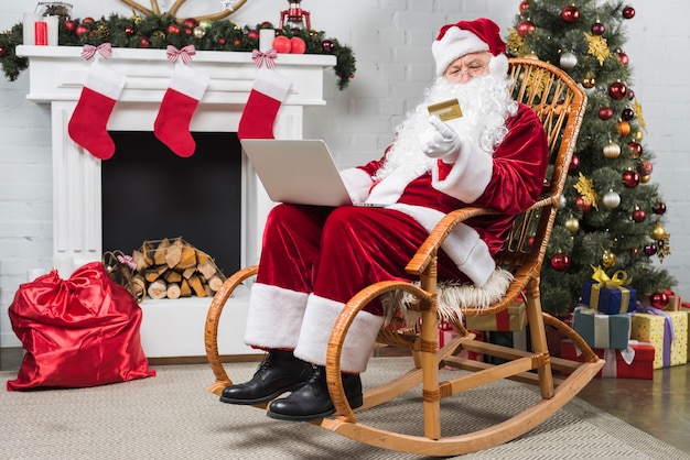 Photo gratuite père noël assis sur une chaise berçante avec ordinateur portable