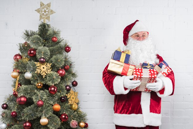 Père Noël avec des cadeaux dans les mains près de sapin de Noël