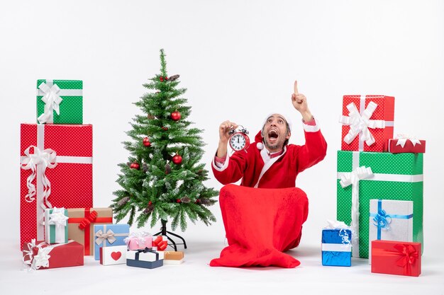 Le père noël excité nerveux pointant au-dessus assis sur le sol et montrant horloge près de cadeaux et arbre de Noël décoré sur fond blanc