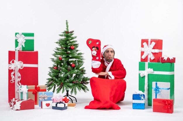 Père Noël triste assis sur le sol et porter des chaussettes de Noël près de cadeaux et arbre du nouvel an décoré sur fond blanc