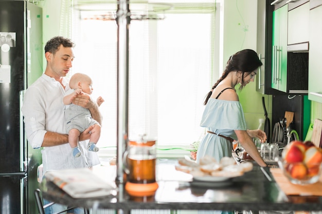 Père portant bébé avec sa femme travaillant dans la cuisine